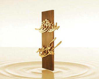May include: A vertical wooden plaque with gold-colored Arabic calligraphy. The plaque is a dark brown color and stands upright against a light background. The calligraphy is ornate and detailed.