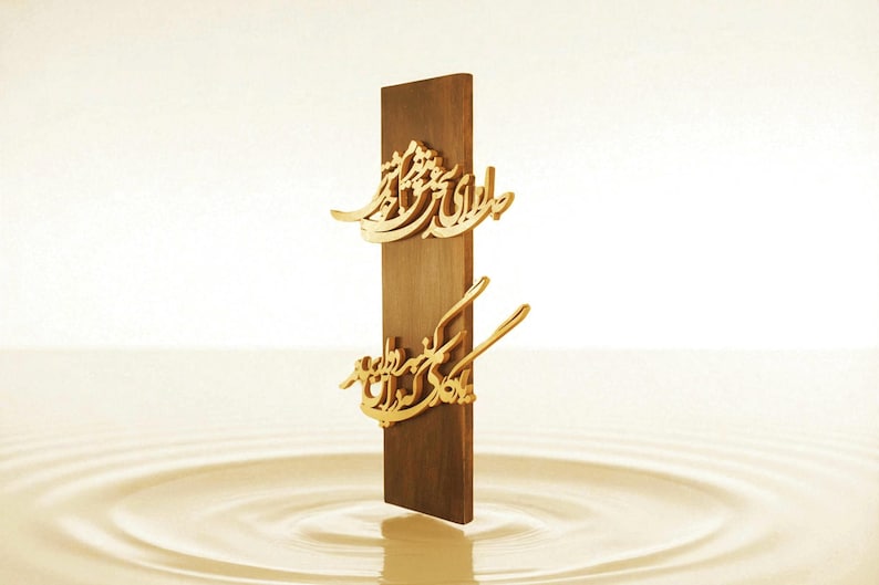 May include: A vertical wooden plaque with gold-colored Arabic calligraphy. The plaque is a dark brown color and stands upright against a light background. The calligraphy is ornate and detailed.