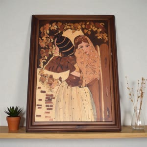 May include: Wooden marquetry art piece in a dark brown frame. The artwork depicts two figures in a garden setting, with a woman in a patterned headscarf and dress. The background features floral and foliage details, with a brick wall and a dark vase.