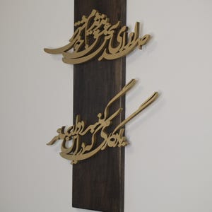 May include: A wooden wall art piece featuring gold-colored calligraphy mounted on a dark brown rectangular base. The calligraphy appears to be in a script style, with two distinct sections of text.
