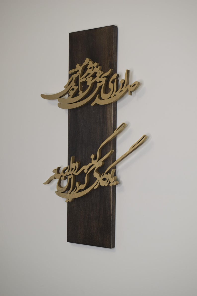May include: A wooden wall art piece featuring gold-colored calligraphy mounted on a dark brown rectangular base. The calligraphy appears to be in a script style, with two distinct sections of text.