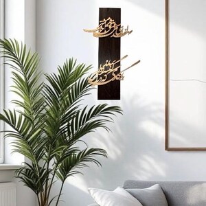 May include: A dark brown wooden wall art piece with gold-colored calligraphy. The artwork is rectangular and mounted vertically. The calligraphy is in an elegant script. The background is a white wall with a plant and a sofa.