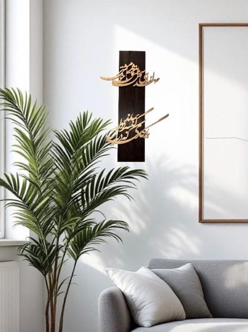 May include: A dark brown wooden wall art piece with gold-colored calligraphy. The artwork is rectangular and mounted vertically. The calligraphy is in an elegant script. The background is a white wall with a plant and a sofa.