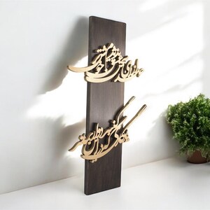 May include: A dark brown wooden wall hanging with gold-colored calligraphy. The calligraphy is in two sections, with the top section having a curved design and the bottom section having a more linear design. The wall hanging is displayed against a white wall.