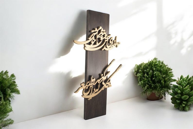 May include: A dark brown wooden wall hanging with gold-colored calligraphy. The calligraphy is in two sections, with the top section having a curved design and the bottom section having a more linear design. The wall hanging is displayed against a white wall.