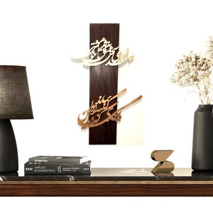 May include: A modern interior scene featuring a dark wooden table with a marble top. On the table are a gray lamp, books, decorative wall art with calligraphy, a black vase with dried flowers, and a gold sculpture.