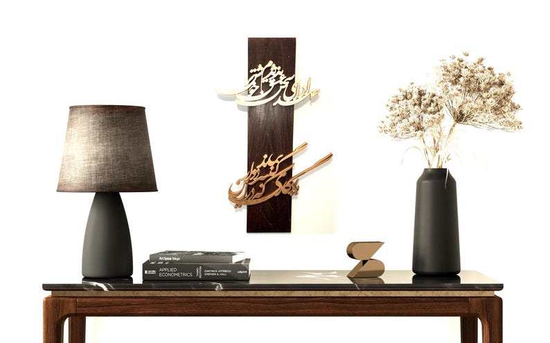 May include: A modern interior scene featuring a dark wooden table with a marble top. On the table are a gray lamp, books, decorative wall art with calligraphy, a black vase with dried flowers, and a gold sculpture.