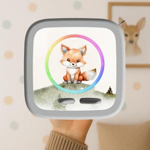 Könnte beinhalten: Ein quadratisches, weißes elektronisches Gerät mit abgerundeten Ecken und einer Aquarellillustration eines kleinen orangefarbenen Fuchses auf einem grünen Hügel. Ein leuchtender, regenbogenfarbener Ring umgibt den Fuchs, darunter befinden sich zwei Tasten.