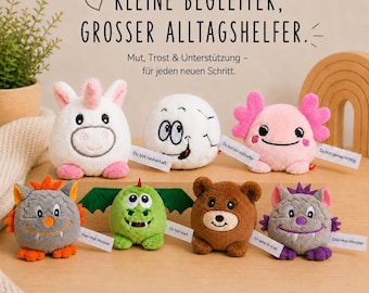 May include: A collection of seven small, round plush toys featuring various characters including a white unicorn, a soccer ball, a pink axolotl, a gray monster, a green dragon, a brown bear, and a purple monster, each with a small white tag.