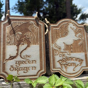 May include: Two wooden plaques featuring a dragon and a horse design. The plaque on the left says "the Green Dragon" and the plaque on the right says "the Prancing Pony". Both plaques are brown with a light brown background and are suspended by string.