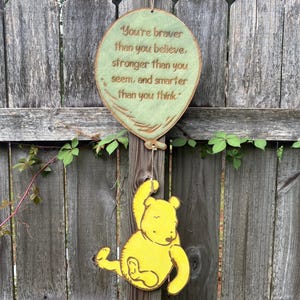 Può includere: Un cartello di legno a forma di palloncino con la scritta "You're braver than you believe, stronger than you seem, and smarter than you think". Un orsetto giallo è appeso sotto il palloncino. Il cartello è appeso a una staccionata di legno.