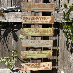 Puede incluir: Un letrero de madera con seis placas rectangulares que representan lugares de Star Wars. Las placas están pintadas en tonos verdes y marrones, con texto e ilustraciones. Los destinos incluyen Kashyyyk, Dagobah y Tatooine. Un juguete de sable de luz es visible en el fondo.