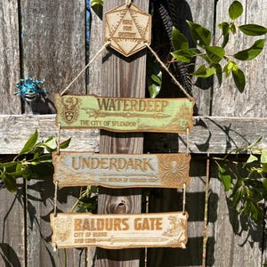 May include: Wooden sign featuring three rectangular plaques with the names "Waterdeep", "Underdark", and "Baldur's Gate" in a fantasy font. The sign is suspended by twine and includes a top piece with the text "Roll for Destiny". The sign is painted in shades of green and brown.