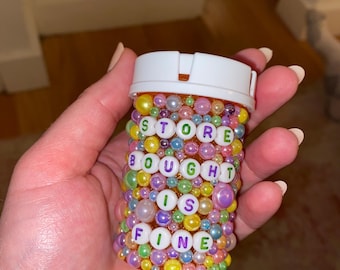 Puede incluir: Un pequeño recipiente de plástico transparente con tapa blanca, decorado con cuentas de colores y letras que forman la frase "STORE BOUGHT IS FINE".