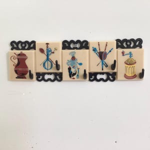 May include: A wall-mounted hook rack featuring five cream-colored square tiles with black scrollwork trim. Each tile displays a colorful kitchen-themed illustration, including a coffee pot, whisk, chef, skewers, and a coffee grinder, with black hooks.
