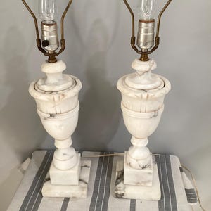 May include: A pair of table lamps crafted from white marble with gray veining. Each lamp features a square base, a pedestal stem, and a fluted urn-shaped body. The lamps include brass-toned metal hardware and a clear light bulb at the top.