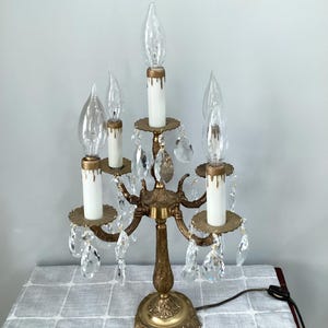 May include: A vintage brass candelabra with five faux candle lights. The candelabra features clear glass flame-shaped bulbs, white candle sleeves, and dangling crystal accents. The base and arms have an ornate design, and the electrical cord is visible.