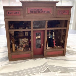 May include: A miniature wooden general store model with a red door and trim. The sign reads "GENERAL MERCHANDISE" and "EST. 1789". The store's interior is visible, filled with tiny items. The model is set on a striped fabric.