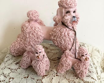 Könnte beinhalten: Ein Set aus drei rosa Pudelfiguren. Die Pudel haben strukturiertes Fell und stehen. Der größte Pudel hat eine Halskette. Die Figuren werden auf einer weißen Spitzentischdecke präsentiert.