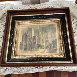 May include: Framed watercolor painting of a European city with tall buildings and a street. The frame is dark wood with a black and gold inner border. The painting has a vintage aesthetic.