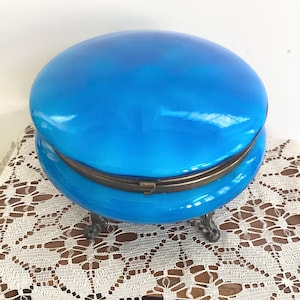 May include: A bright blue, oval-shaped trinket box with a hinged lid and ornate metal feet. The box has a glossy finish and a brass-colored metal clasp. The box is sitting on a white lace tablecloth.