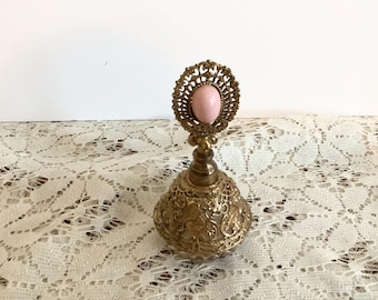 May include: A gold-toned perfume bottle with a detailed filigree design and a pink glass stopper. The bottle is sitting on a white lace surface.