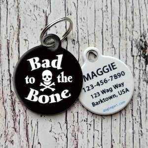 May include: A round black pet ID tag featuring a white skull and crossbones graphic with the text Bad to the Bone. A second white tag shows the name MAGGIE, the phone number 123-456-7890, the address 123 Wag Way, Barktown, USA, and dogtagart.com.