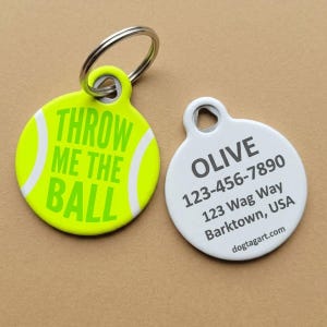 May include: Two round pet tags. One is neon yellow with white curved lines resembling a tennis ball and the text THROW ME THE BALL. The other is white with the text OLIVE, 123-456-7890, 123 Wag Way, Barktown, USA, and dogtagart.com. A metal ring is attached.