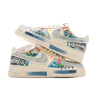 Puede incluir: Un par de zapatillas deportivas blanquecinas con detalles de mezclilla azul y trazos de pintura coloridos. Las zapatillas presentan las palabras "HAPPY" y "So" con un gr&aacute;fico de carita sonriente. Las suelas son blanquecinas con un dise&ntilde;o moteado.