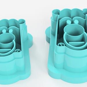 May include: Two turquoise plastic cookie cutters. Each cutter has a rounded base with a scalloped edge, a central circular cup, and a series of vertical and curved supports. The cutters are designed to create a bear shape.