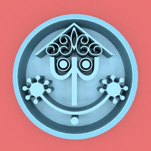 May include: A light blue circular design with a house-like top, two eyes, and a smiling face. The design has two snowflake-like shapes on either side of the face. The background is a solid coral color.