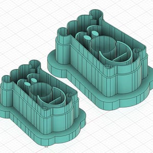 May include: Two teal-colored cookie cutters in the shape of a baby's foot. The cutters have a rectangular base with a rounded top and a detailed interior design. The cutters are made of plastic and are designed for baking.