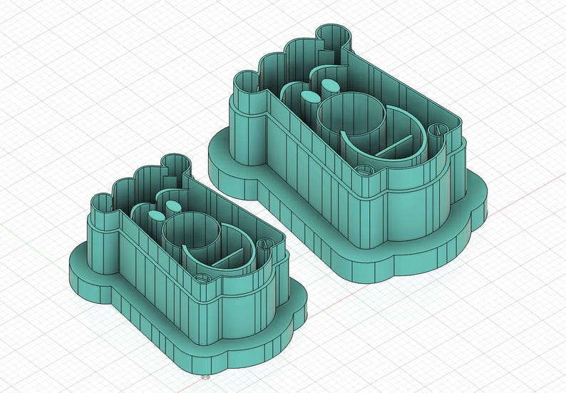 May include: Two teal-colored cookie cutters in the shape of a baby's foot. The cutters have a rectangular base with a rounded top and a detailed interior design. The cutters are made of plastic and are designed for baking.