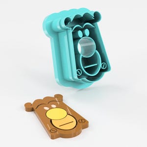 May include: A two-piece cookie cutter set. The larger piece is teal and shaped like a face with a mustache and a circular nose. The smaller piece is brown with a yellow circle and a horizontal line, resembling a face.