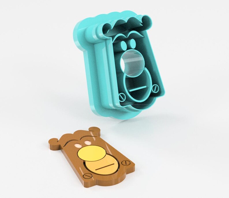 May include: A two-piece cookie cutter set. The larger piece is teal and shaped like a face with a mustache and a circular nose. The smaller piece is brown with a yellow circle and a horizontal line, resembling a face.