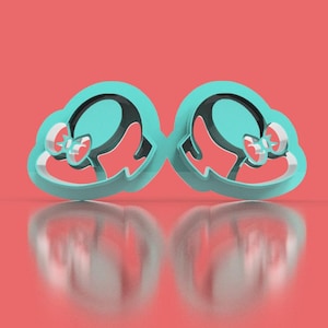 May include: Two matching teal cookie cutters in the shape of a hat with a bow. The interior of the hat is black, and the bow is red. The cutters are on a reflective surface with a coral background.