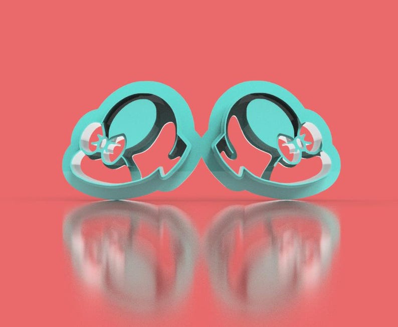 May include: Two matching teal cookie cutters in the shape of a hat with a bow. The interior of the hat is black, and the bow is red. The cutters are on a reflective surface with a coral background.