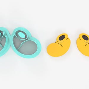 May include: A set of two teal-colored cookie cutters in a heart shape, and two yellow cookie cutters shaped like cartoon shoes. The cookie cutters are made of plastic and are on a white background.
