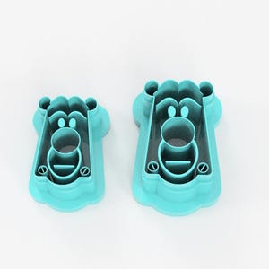 May include: Two turquoise plastic cookie cutters in the shape of a bear's face. The cutters have a detailed design with a round nose, eyes, and a mouth. The cutters are different sizes, with the larger one on the right.