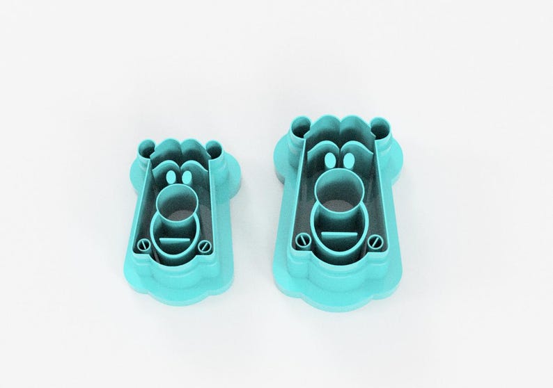 May include: Two turquoise plastic cookie cutters in the shape of a bear's face. The cutters have a detailed design with a round nose, eyes, and a mouth. The cutters are different sizes, with the larger one on the right.