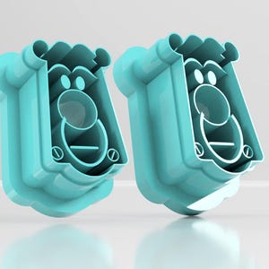 May include: Two turquoise-colored, cartoon-like face-shaped cookie cutters. Each cutter has a rounded top, a circular nose, and a curved mouth. The design includes eye and mouth details, and a decorative bottom edge.