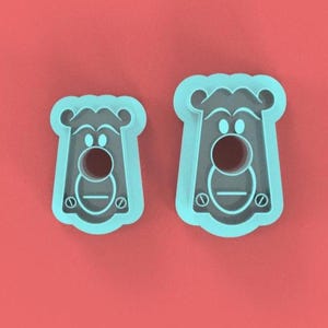 May include: Two light blue bear-shaped cookie cutters on a red background. The cutters have a circular hole for the bear's nose and a horizontal line for the mouth. The bear's face has two small circles for eyes and a rounded top.