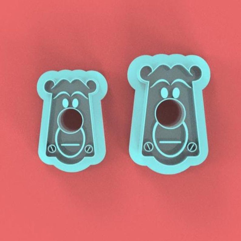 May include: Two light blue bear-shaped cookie cutters on a red background. The cutters have a circular hole for the bear's nose and a horizontal line for the mouth. The bear's face has two small circles for eyes and a rounded top.