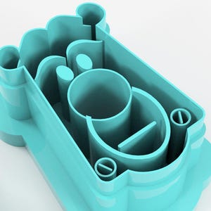 May include: A light teal-colored cookie cutter with a complex design. The cutter features a rectangular shape with rounded edges and several internal cutouts, including a central cylinder and a small, circular detail.