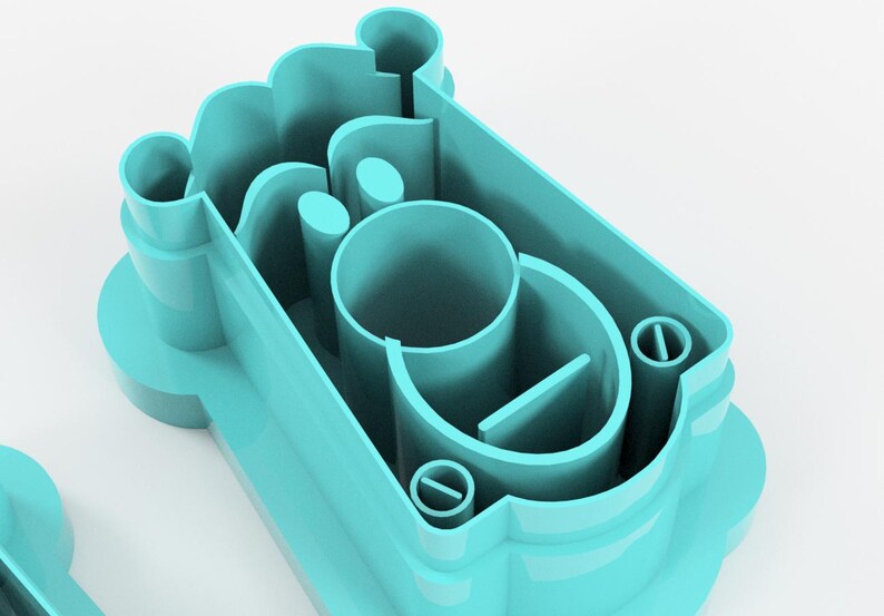 May include: A light teal-colored cookie cutter with a complex design. The cutter features a rectangular shape with rounded edges and several internal cutouts, including a central cylinder and a small, circular detail.