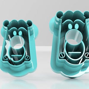 May include: Two turquoise plastic cookie cutters in the shape of a bear's face. The cutters have a circular opening for the mouth and two smaller circles for the eyes. The bear's face has a rounded top and a scalloped bottom.