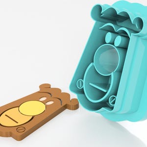 May include: A two-part cookie cutter set. The brown cookie has a yellow circle and a line. The blue mold has a matching shape. The set is designed to cut out a bear-shaped cookie.