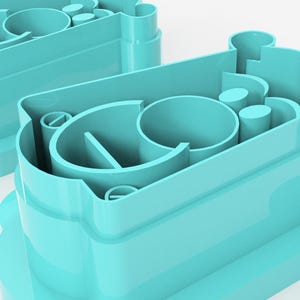 May include: Two turquoise plastic cookie cutters. Each cutter has a rounded base with a scalloped edge and a complex interior design featuring curved and cylindrical shapes. The cutters are designed for baking.