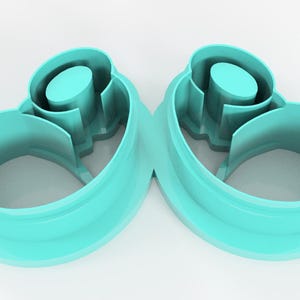 May include: Two teal-colored, abstract-shaped cookie cutters. Each cutter has a rounded, open base that curves upwards, with a central cylindrical element and interior cutouts. The cutters are made of plastic and are set against a white background.