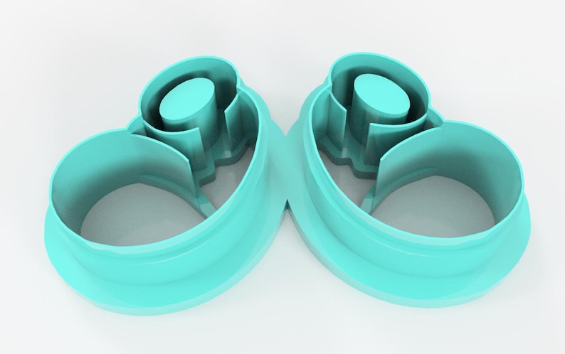 May include: Two teal-colored, abstract-shaped cookie cutters. Each cutter has a rounded, open base that curves upwards, with a central cylindrical element and interior cutouts. The cutters are made of plastic and are set against a white background.
